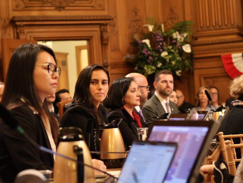 A group of people in formal attire sit at a conference table with laptops and coffee carafes, discussing SF family shelter stays 2025, in a wood-paneled room during a meeting.