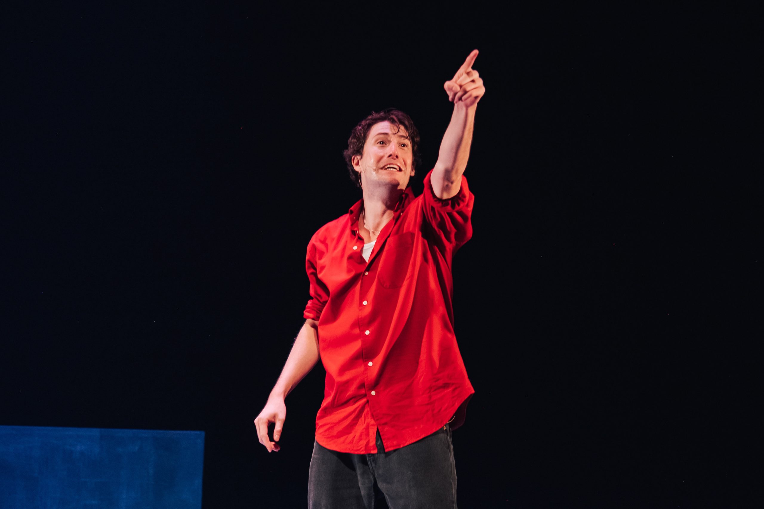 A person in a red shirt stands on stage, pointing upward with their right hand against a dark background.