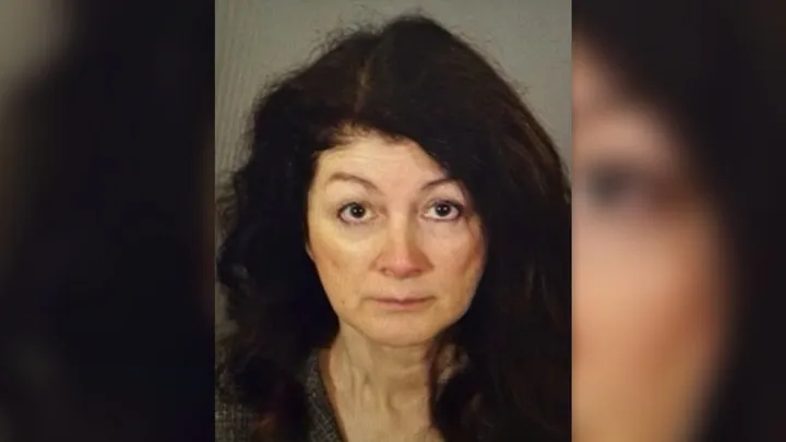 A woman with long dark hair and a neutral expression looks directly at the camera against a plain background.