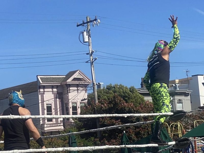 Two masked wrestlers stand in a ring outdoors; one in green gear reaches upward while the other in blue watches. Houses and power lines frame the scene, offering a buena vista of this lively match.