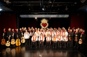 A large musical group poses on stage with various string instruments, all wearing coordinated outfits, in front of a backdrop featuring a heart-shaped decoration.