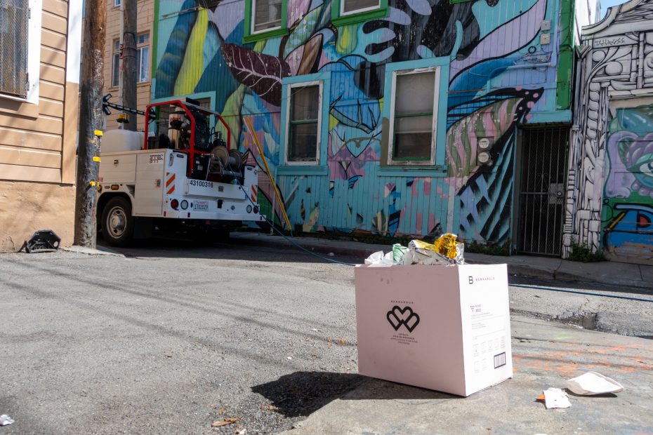 Utility truck parked in alley near a colorful mural-covered building, with a box of trash in the foreground.
