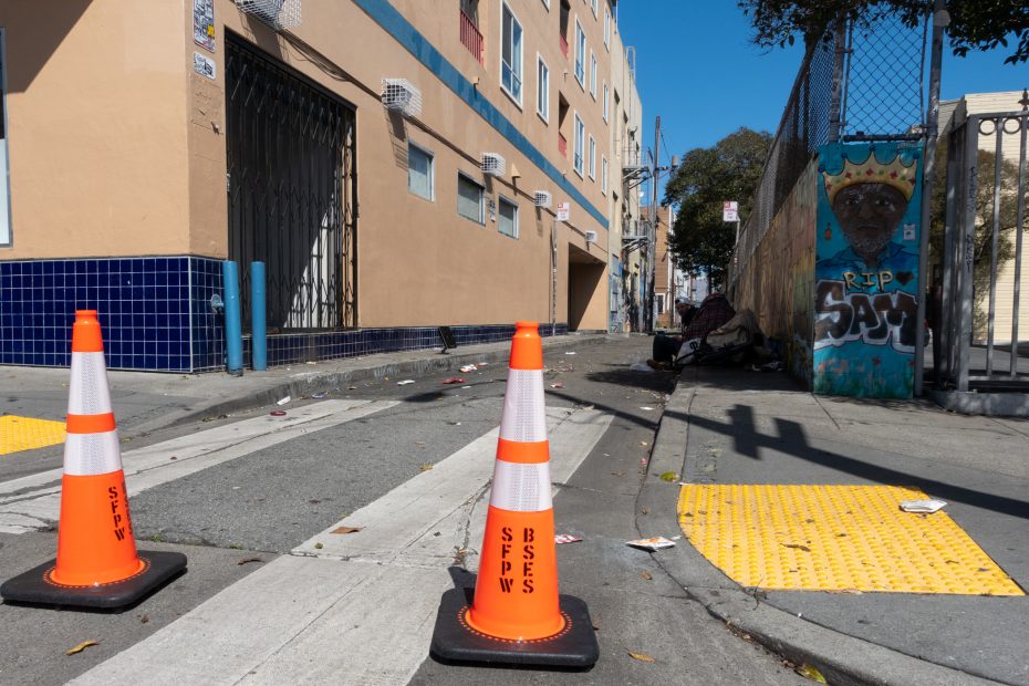 Two traffic cones on a pedestrian crossing near a building. An alley with litter, a fence, and a street art mural is visible.