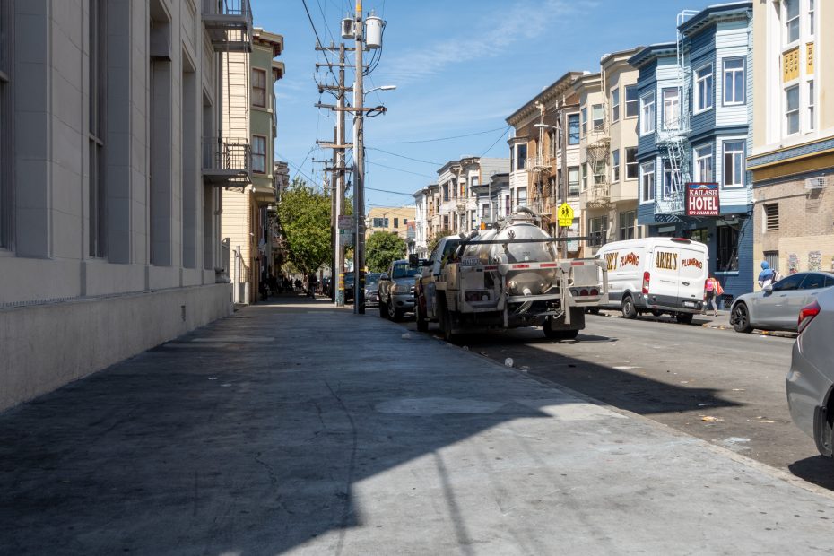 A city street lined with parked cars and colorful buildings under a clear blue sky. A truck and a van are visible in the foreground.