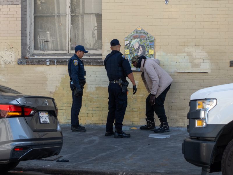 Two police officers interact with a person on a city sidewalk. The person is bent over, facing the officers. Cars are parked nearby, and a mural is on the wall behind them.