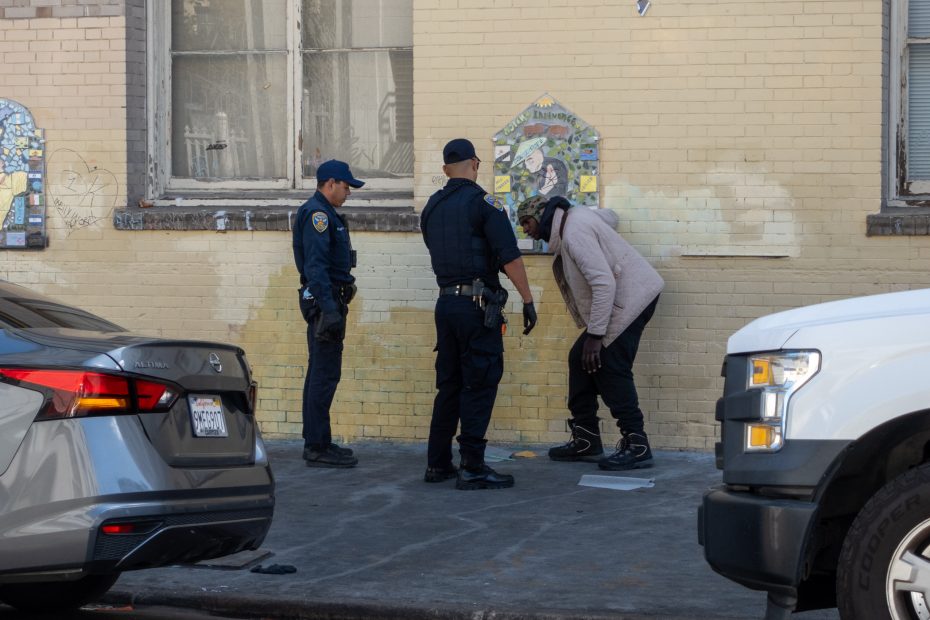 Two police officers interact with a person on a city sidewalk. The person is bent over, facing the officers. Cars are parked nearby, and a mural is on the wall behind them.