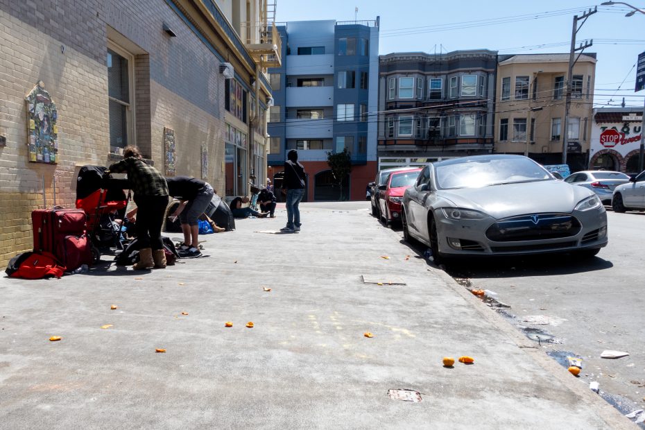 People sorting through belongings on a sidewalk near a Tesla parked on the street; scattered trash and buildings in the background.