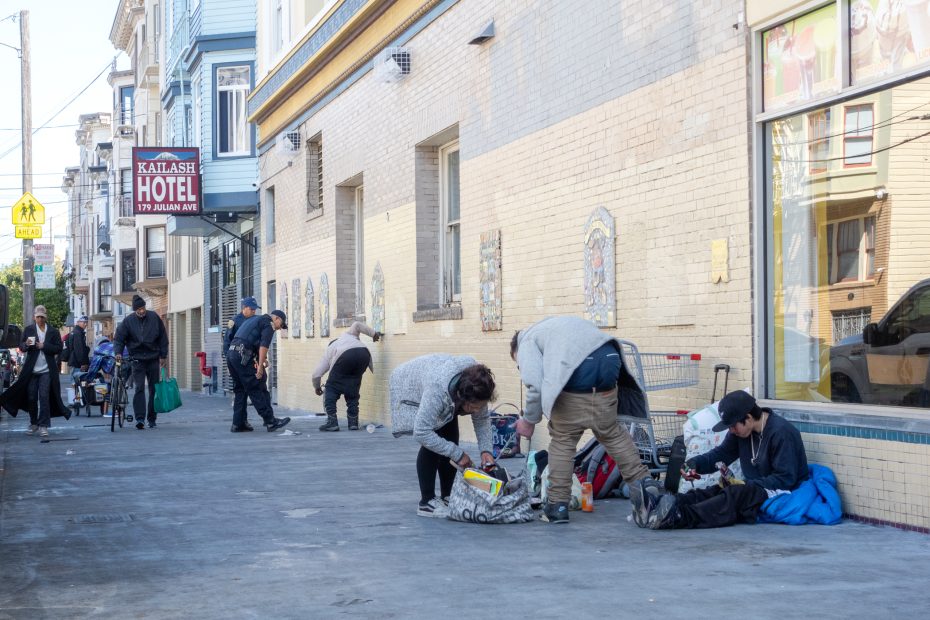 People are gathered on a sidewalk near a brick building with a "Kailash Hotel" sign. Some are crouching and organizing belongings, while others stand or walk by.
