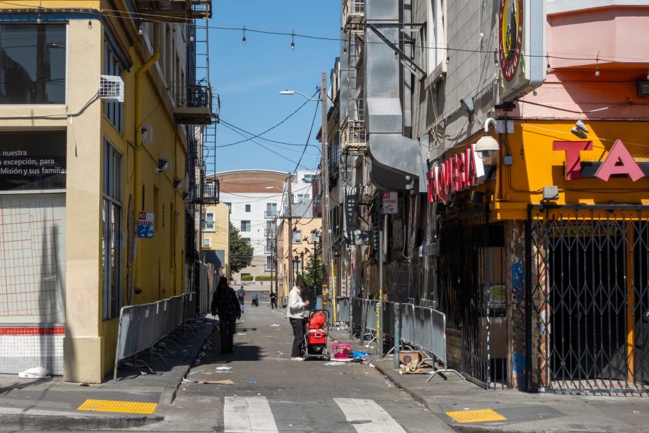 Narrow urban alleyway with scattered debris, buildings on either side, and a few people walking. Yellow and orange storefronts visible. String lights hang overhead.