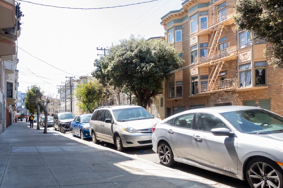 A city street with parked cars, a sidewalk, and multi-story residential buildings with fire escapes. Trees line the sidewalk. The scene is in daylight.