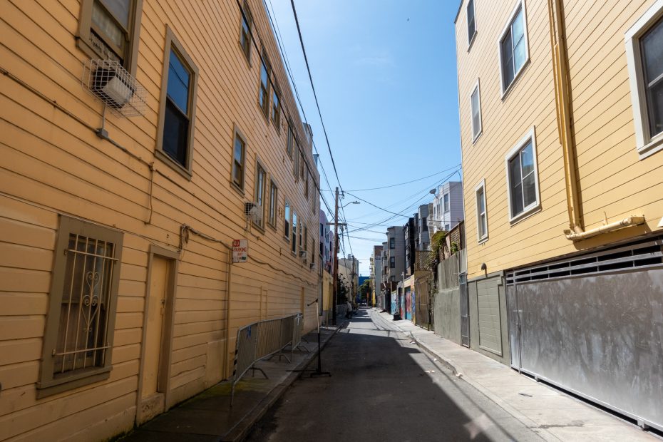 Narrow urban alley with yellow buildings on both sides, overhead utility lines, and scattered barricades. Clear blue sky above.