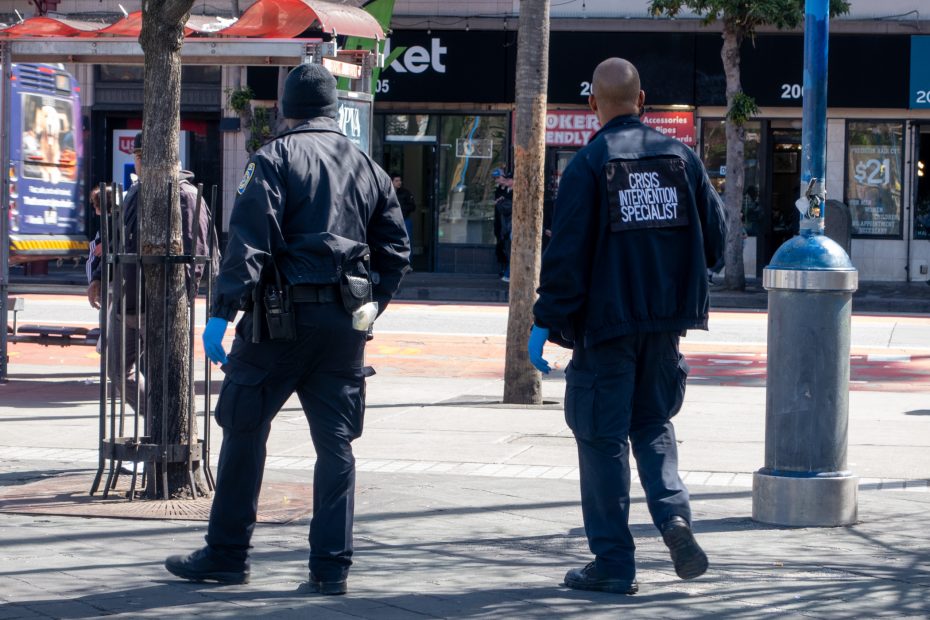 Two men in uniforms, one labeled "Crisis Intervention Specialist," walk on a city street wearing gloves.
