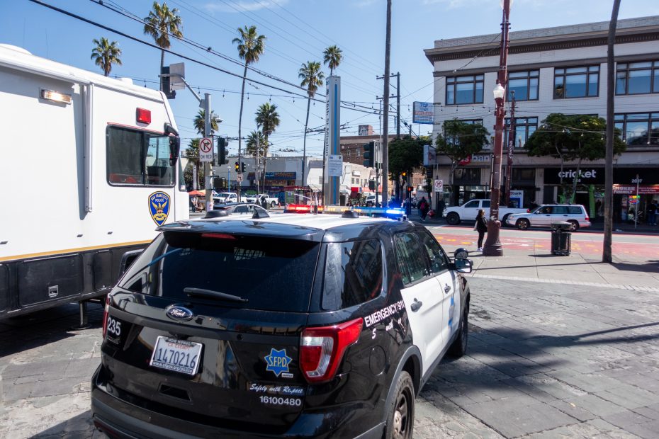 Police vehicles with lights on at a city intersection. Palm trees, buildings, and a few people in the background.
