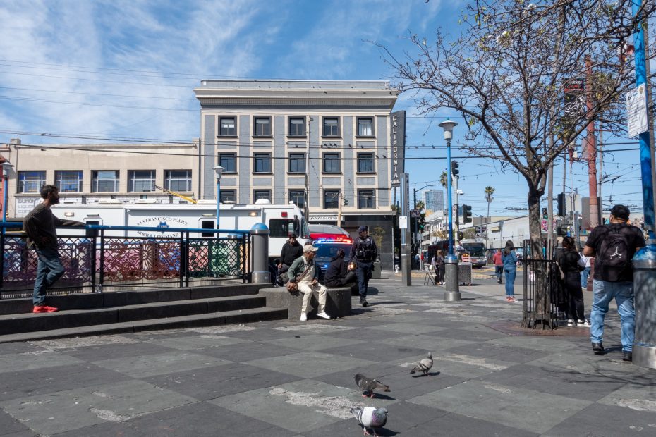 Urban scene with people walking and sitting near a city square. Pigeons on the pavement, a building, and parked emergency vehicles in the background under a blue sky.