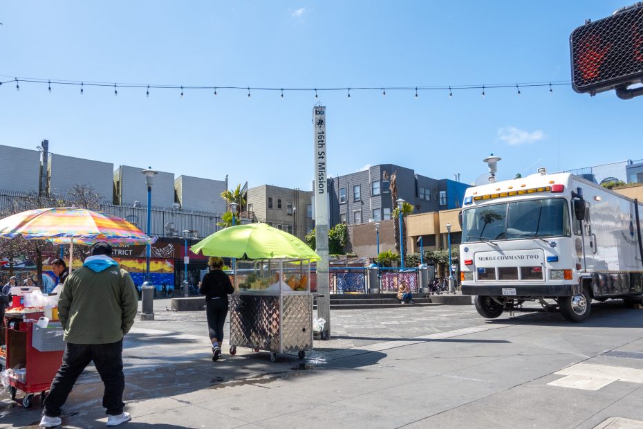 Street scene with food vendors, a truck, and pedestrians in a plaza under string lights. Buildings are in the background on a sunny day.