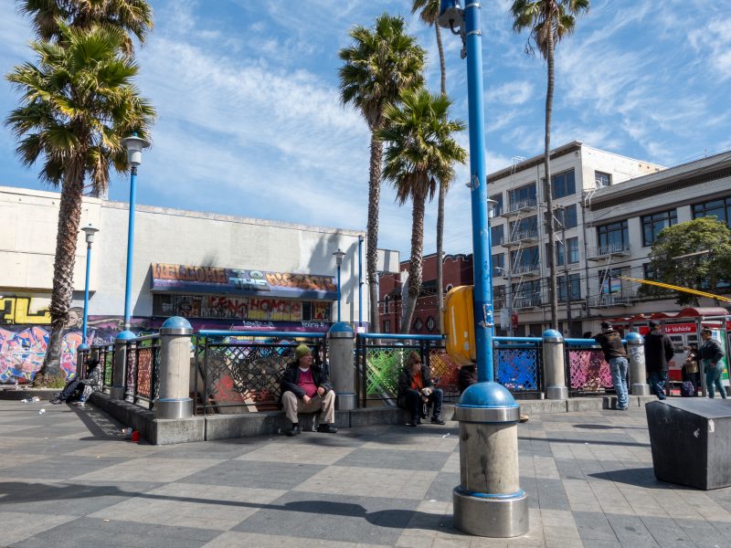 Urban plaza with people sitting and standing, surrounded by palm trees and buildings. Graffiti-covered walls and a sunny sky are visible.
