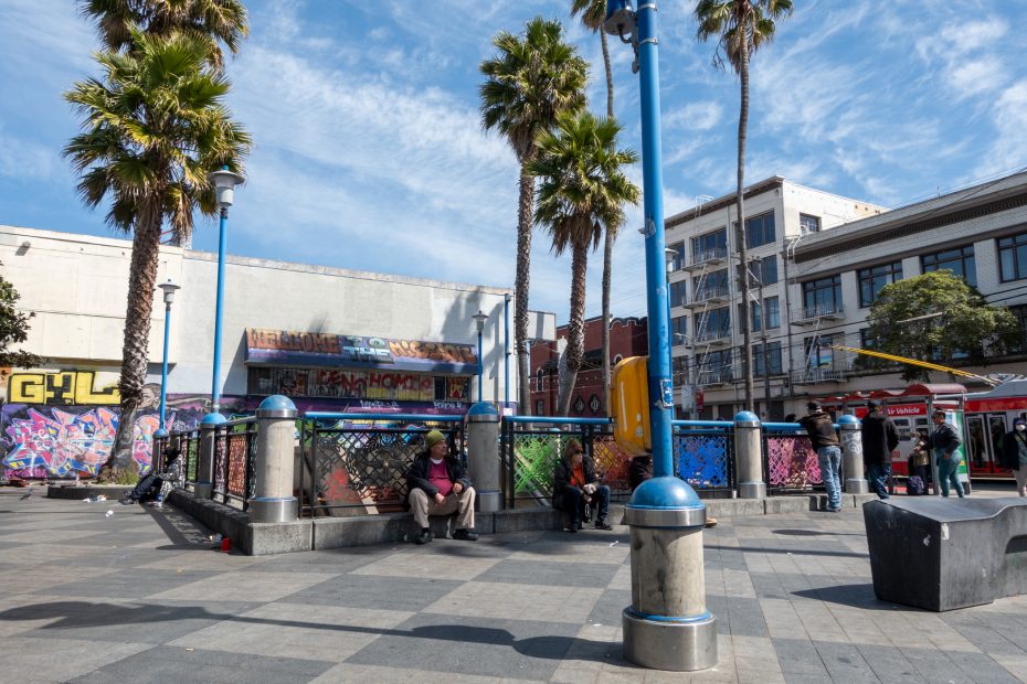 Urban plaza with people sitting and standing, surrounded by palm trees and buildings. Graffiti-covered walls and a sunny sky are visible.