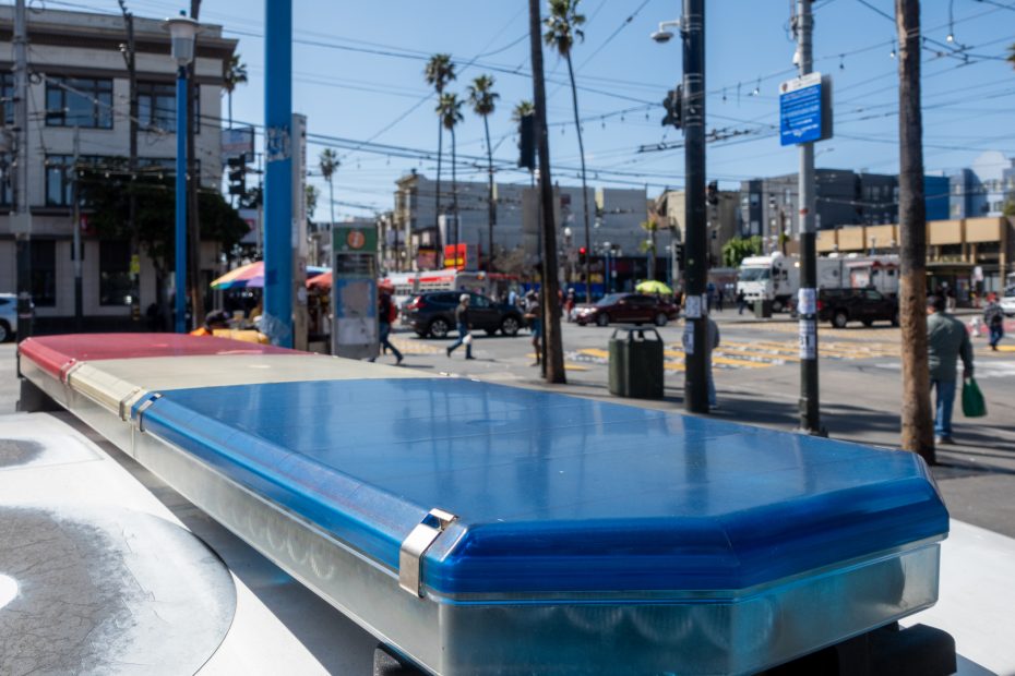 View of a city street with a blue emergency vehicle light bar in the foreground, buildings, palm trees, and traffic in the background.
