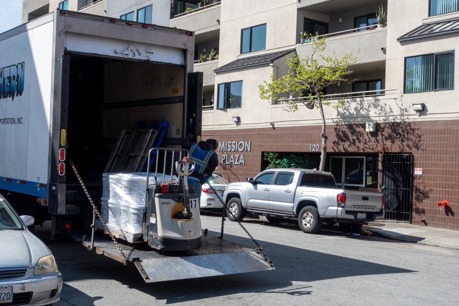 Person operates a pallet jack to load or unload items from a truck on a city street near an apartment building.