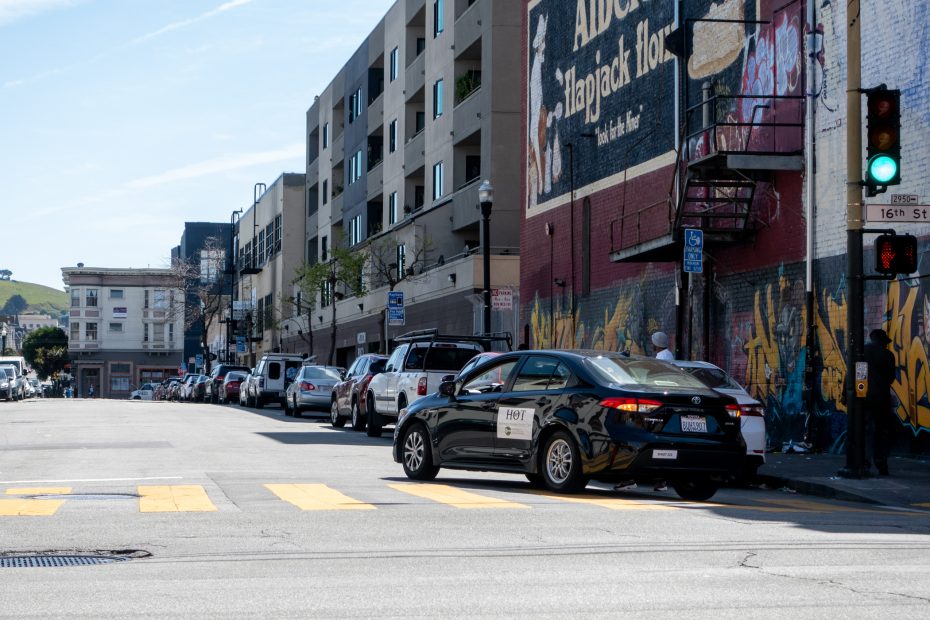 A black car waits at a red light on a city street. Buildings and parked cars line the road. A mural and traffic signs are visible in the background.