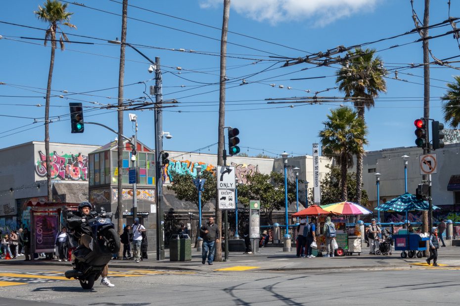A motorcyclist performs a wheelie on a city street corner. Graffiti-covered buildings, palm trees, street signs, and a small crowd feature in the urban backdrop.