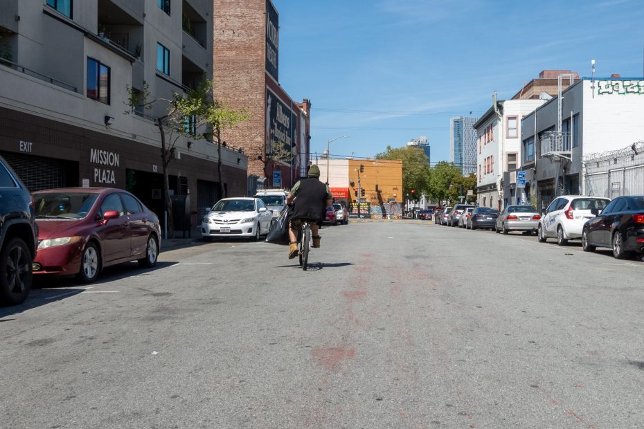 Person in black jacket riding a bicycle down an empty city street with parked cars on both sides, under a clear blue sky.
