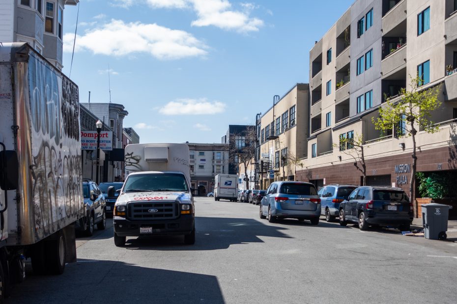 City street with parked vehicles, including a graffiti-covered truck. Modern apartment buildings line both sides. Clear blue sky with a few clouds overhead.