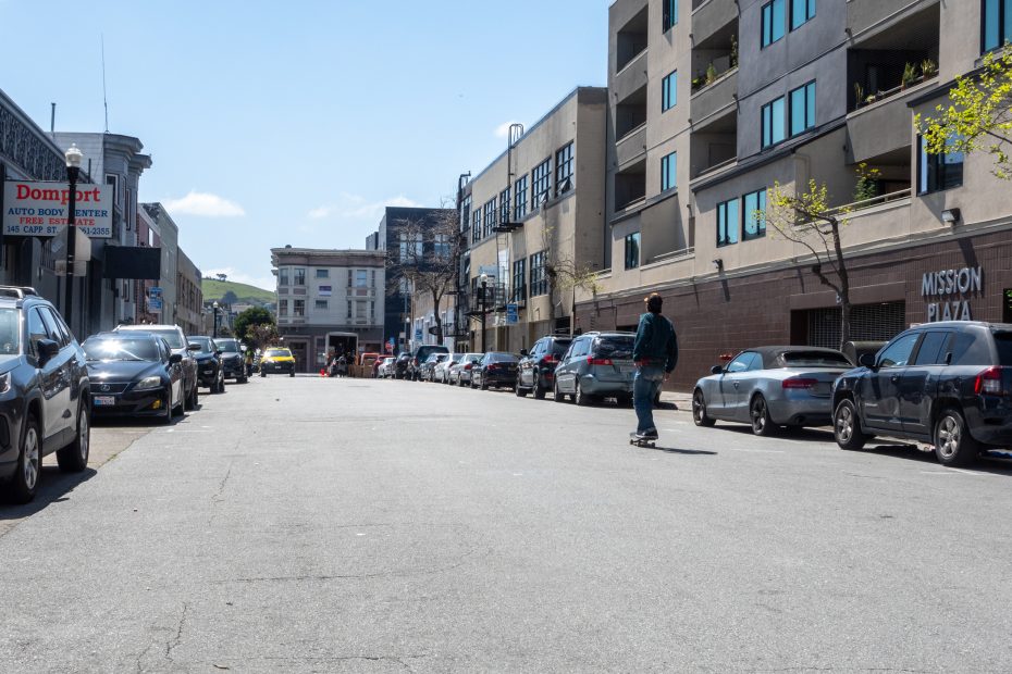 A person skateboards down an urban street lined with parked cars and buildings on a clear day.