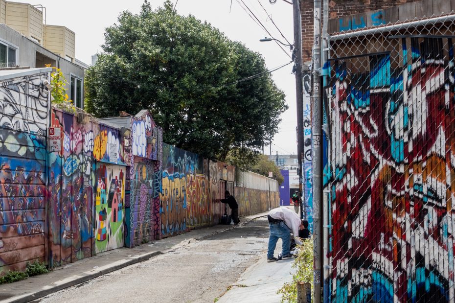 A narrow alleyway with two people and walls covered in colorful graffiti. One person is crouched down, and another stands near an artwork. A large tree is visible in the background.