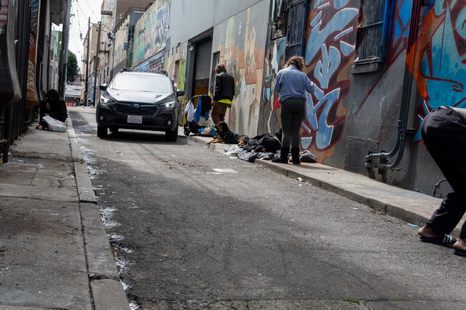 Narrow alley with street art on the walls, several people sitting or standing, and a parked car. Trash and debris are scattered along the sides.