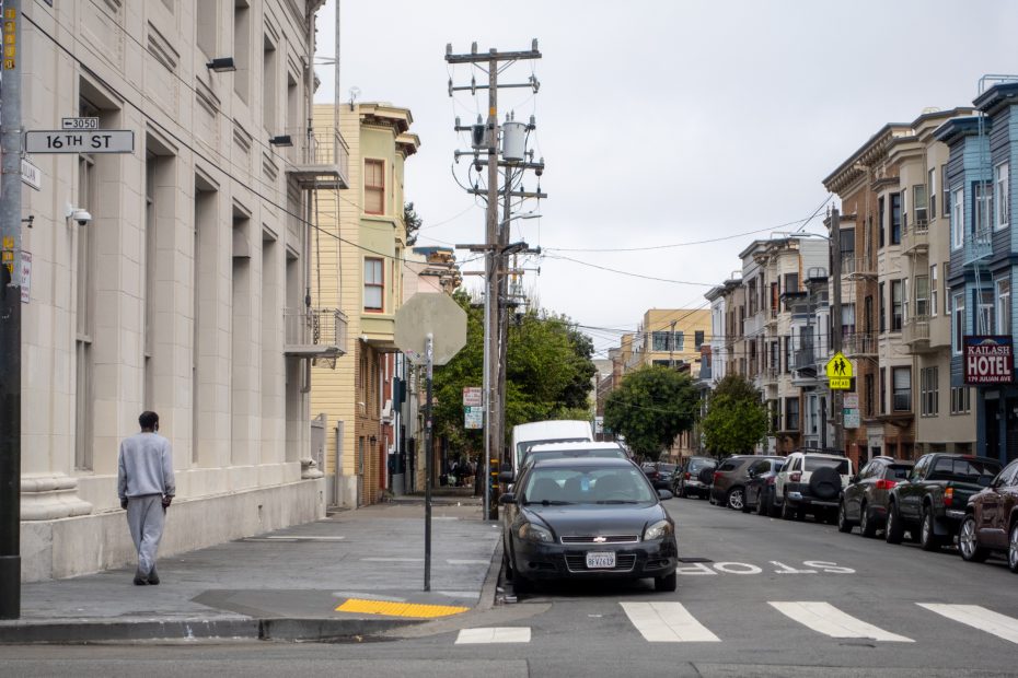 Street view with parked cars, trees, and buildings on both sides. A person walks on the sidewalk. A sign reads "16th St". Overcast sky.