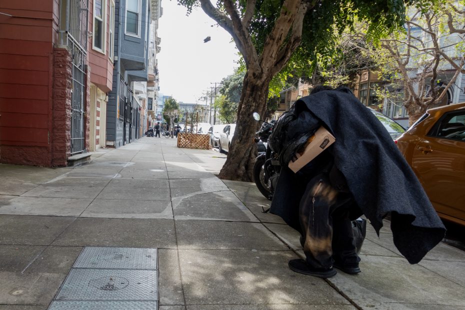 Person walking on a city sidewalk, carrying items under a blanket. Trees and parked cars line the street.