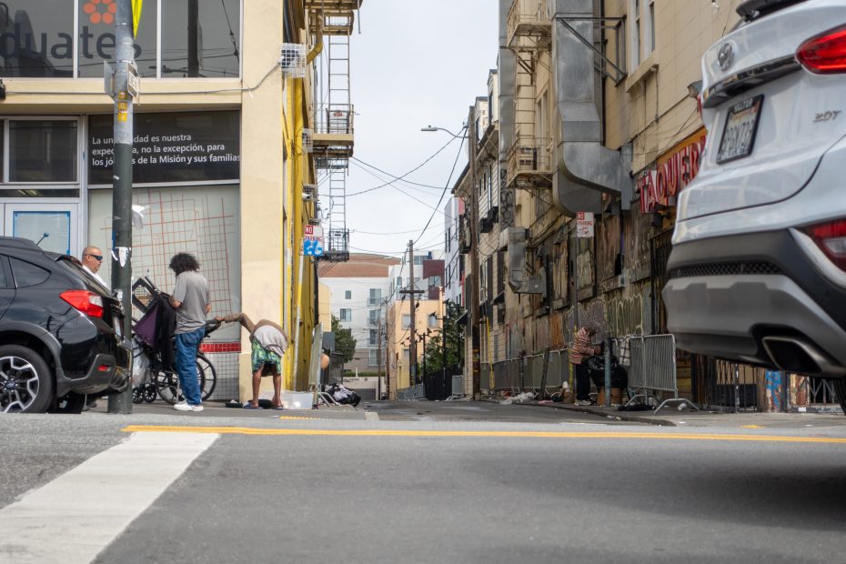 Street scene with people on the sidewalk, parked cars, and buildings. One person bends down near bags. Urban environment with visible wires and utility poles.
