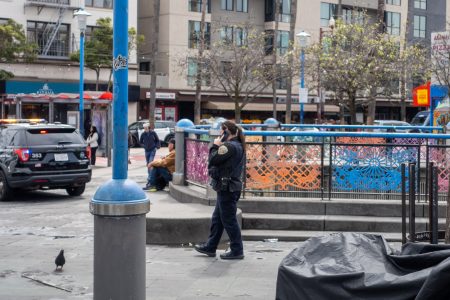 A police officer talks on a phone while walking past colorful fence panels in an urban area, with buildings, parked police cars, and a pigeon nearby.