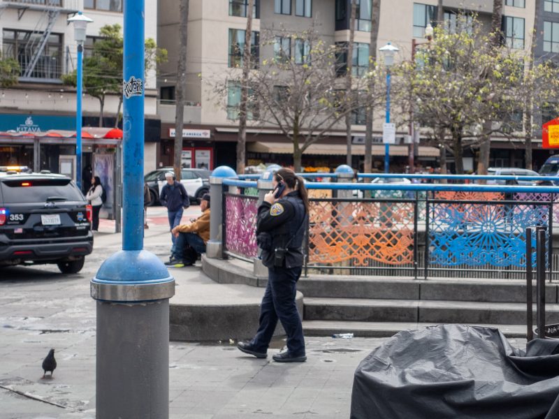 A police officer talks on a phone while walking past colorful fence panels in an urban area, with buildings, parked police cars, and a pigeon nearby.