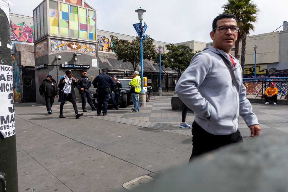 A street scene with people walking, police officers talking near a patrol car, and a graffiti-covered building in the background. A man in a gray hoodie walks in the foreground.