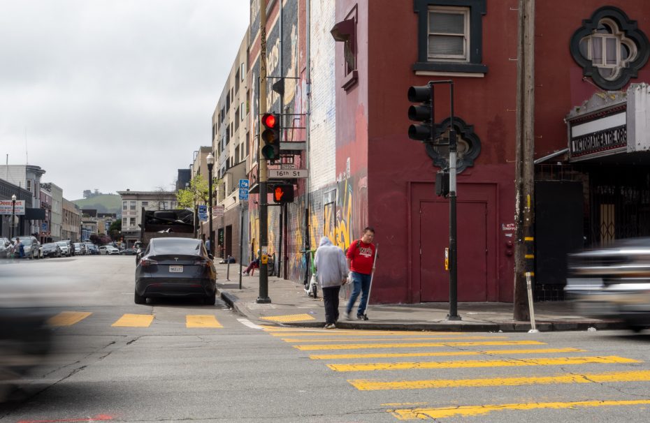 Urban street corner with a red building and mural. Two people cross at a yellow crosswalk while cars wait and pass by. A traffic light shows red.
