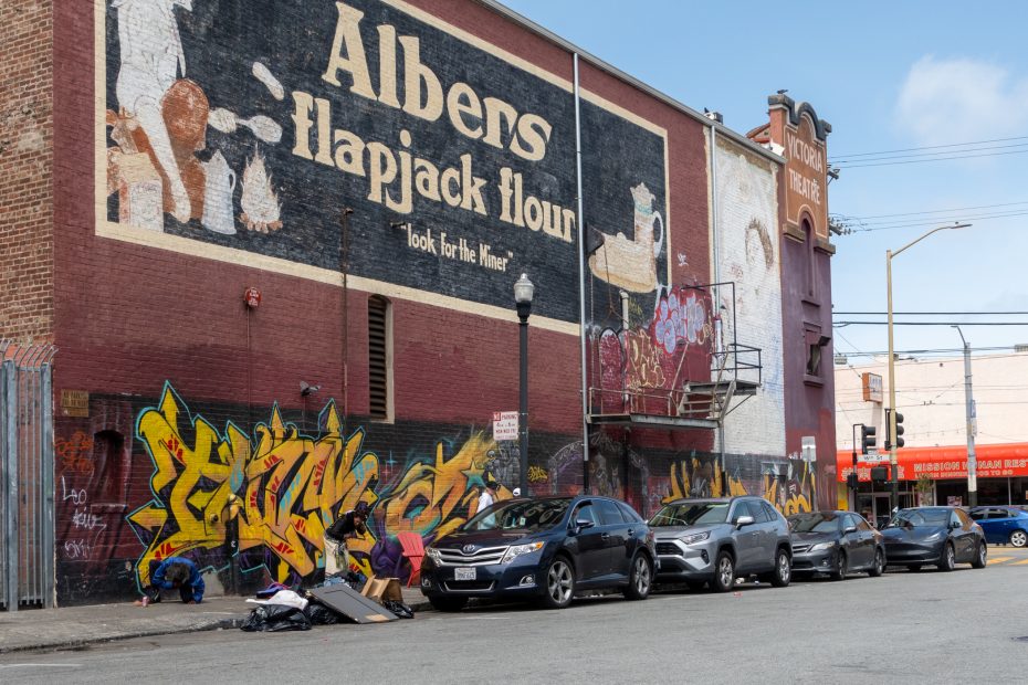 Street view of a row of parked cars in front of a brick building with graffiti and a large vintage "Albens Flapjack Flour" mural. People are gathered on the sidewalk near the building.