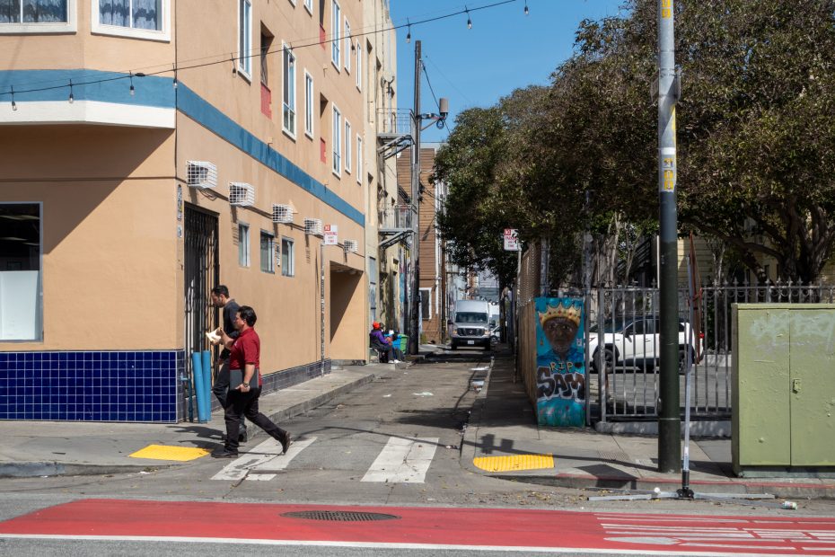 Street scene with people walking past buildings and a mural of a bear on a wall. Trees and a parked vehicle are visible in the background.