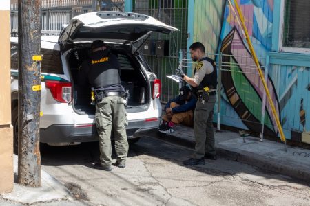 Two officers from a sheriff's department are handling paperwork and gear from a vehicle. Nearby, a seated individual is wearing a hooded jacket, with colorful wall art in the background.
