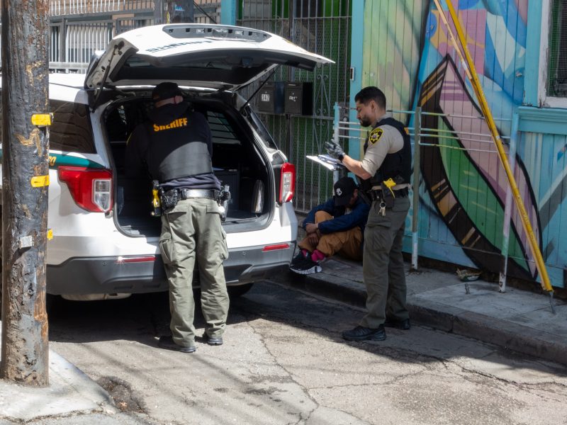 Two officers from a sheriff's department are handling paperwork and gear from a vehicle. Nearby, a seated individual is wearing a hooded jacket, with colorful wall art in the background.