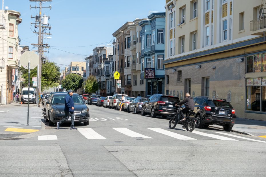 A city street with a crosswalk. A person crosses the road, while another rides a bicycle. Parked cars line the street beside apartment buildings under a clear sky.