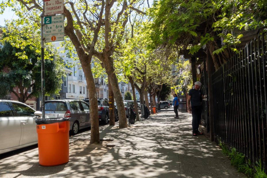 A sunny sidewalk with parked cars on the left and a person standing near a fence on the right. Trees cast dappled shadows, and there's an orange trash bin in the foreground.