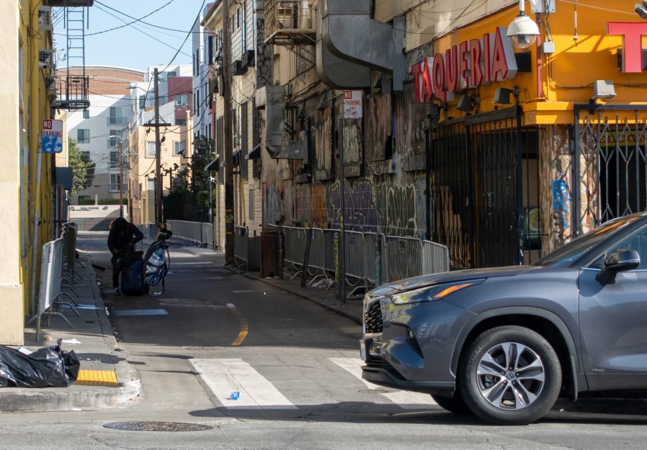 A person collects belongings in an alley beside a graffiti-covered wall, with a parked SUV and a taqueria nearby.