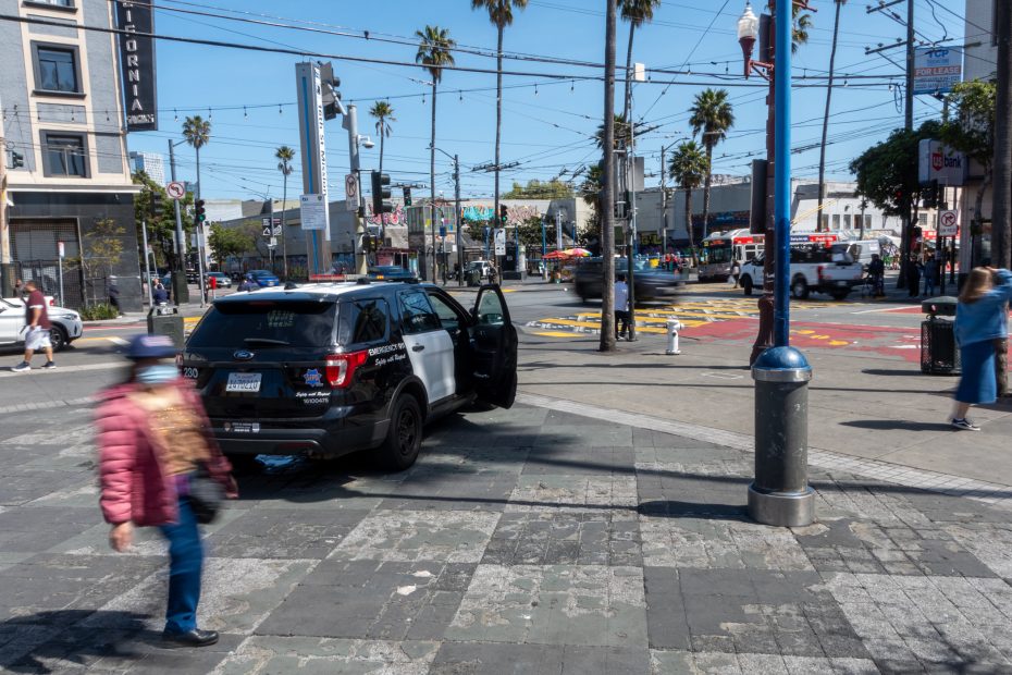 Police SUV parked on a city sidewalk with open driver's door, surrounded by buildings, palm trees, and pedestrians in motion.