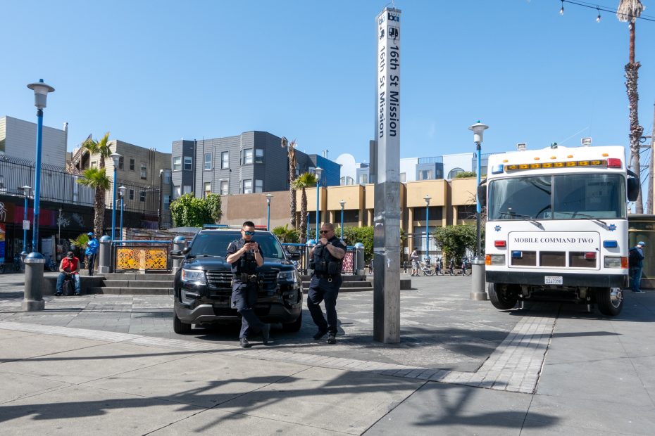 Two police officers stand near a police vehicle at the 16th St Mission station, with a Mobile Command Two truck nearby and several people in the background.