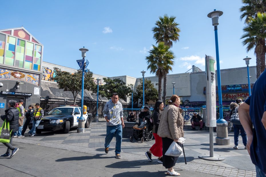 People walking and sitting near a street lined with tall lampposts and palm trees. Buildings in the background have colorful murals. A parked taxi and two seated police officers are visible.