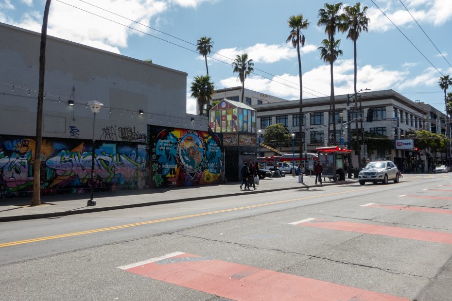 Street view with a building showcasing colorful graffiti, palm trees, and a bright house-shaped structure on the roof. Pedestrians and cars are visible under a partly cloudy sky.
