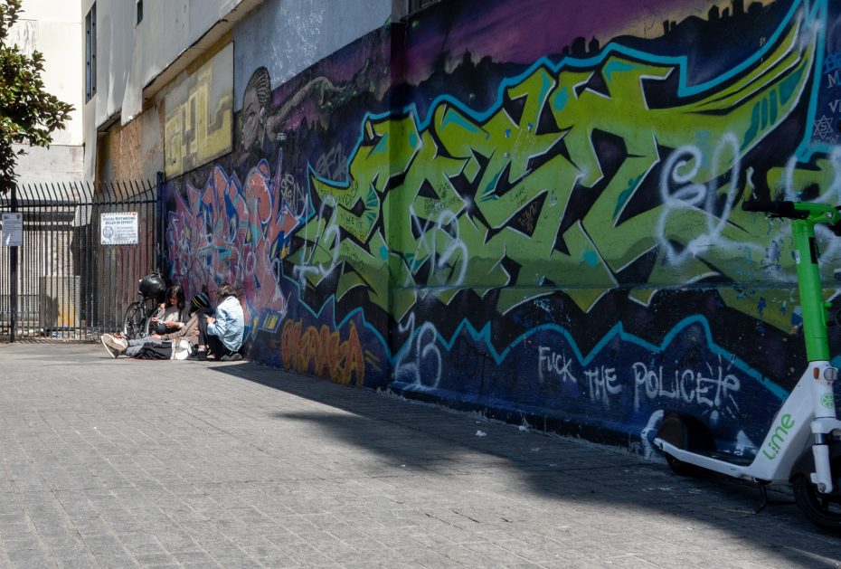Two people sitting on a sidewalk beside colorful graffiti-covered walls, with a green electric scooter in the foreground.