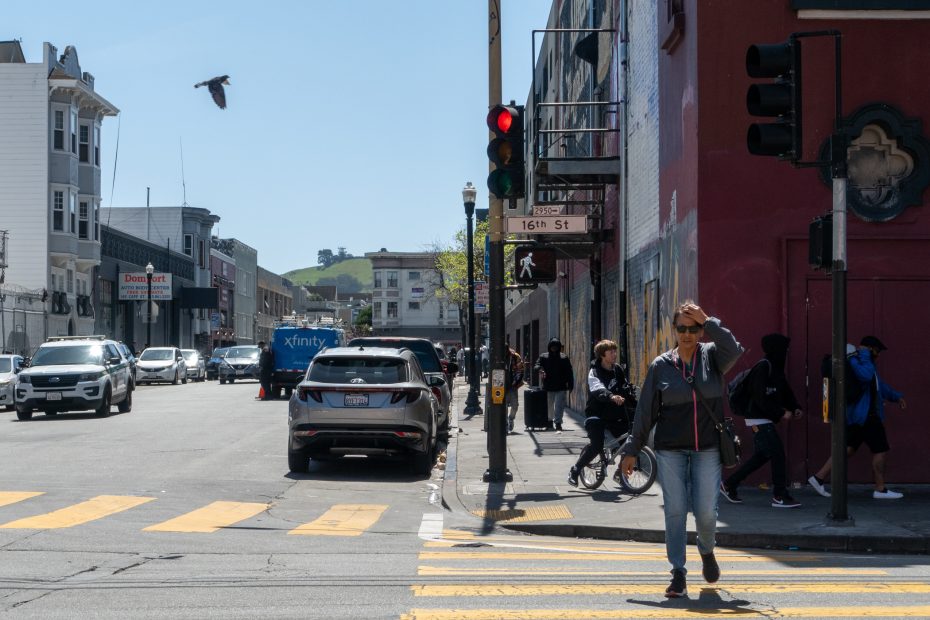 A city street with people walking, cars at a red light, and a bird flying overhead. Buildings and a light pole are visible.